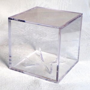 May include: A clear acrylic display cube with a removable lid. Inside, a clear plastic stand is visible, designed to hold a spherical object. The cube is designed to showcase collectibles.