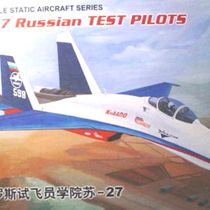 May include: A white, blue, and red SU-27 Russian Test Pilots model airplane with the text "КНААПО" and "598" on the side. The model is on a brown dirt runway.