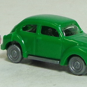 May include: A green toy car with a black figure standing next to it. The car has a rounded shape and a silver grill. The figure is wearing a black jacket and pants.