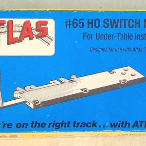 Puede incluir: Una caja azul y amarilla con el texto "ATLAS #65 HO SWITCH MACHINE For Under-Table Installation Designed for use with Atlas Turnouts You're on the right track... with ATLAS!" La caja muestra una máquina de cambio de tren modelo.