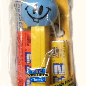 May include: A blue and yellow PEZ dispenser with a smiling face on the top. The dispenser is packaged in a clear plastic bag with the words "Candy & Dispenser" printed on the front. The bag also includes the text "Interactive Classic" and "Enjoy other PEZ Candy Flavors. Collect different PEZ Dispensers."