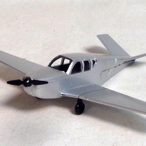 Puede incluir: Un modelo de avión de color plateado con una hélice y tren de aterrizaje negros. El avión tiene un ala individual a cada lado y una aleta de cola. Las ventanas de la cabina son transparentes. El modelo es una réplica en miniatura de una aeronave pequeña.