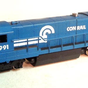 May include: A blue model train locomotive with the words "CONRAIL" and "1991" printed on the side. The locomotive has a white logo with a stylized "C" on the side.