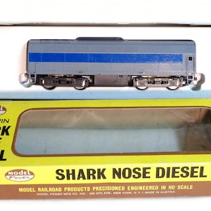 May include: A model train, a Baldwin Shark Nose Diesel, is displayed in its original box. The train is gray with a blue stripe and black wheels. The box is yellow and brown with the text "SHARK NOSE DIESEL".