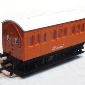 May include: Orange toy train car with a white roof and a smiling face. The car has several windows and the word "Clarabel" printed on the side.