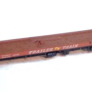 Puede incluir: Un vagón de tren modelo marrón con las palabras "TRAILER TRAIN" impresas en el lateral. El vagón tiene una plataforma plana y está hecho de metal.