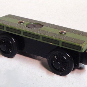 Puede incluir: Un vagón de tren de juguete de madera negro con una plataforma verde. La plataforma tiene un patrón de cuadrícula y un círculo en el centro.