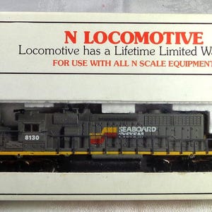 May include: A model train locomotive in a box. The locomotive is gray with yellow and red accents, and the box has the Bachmann logo and text that reads "N Locomotive" and "For use with all N scale equipment."