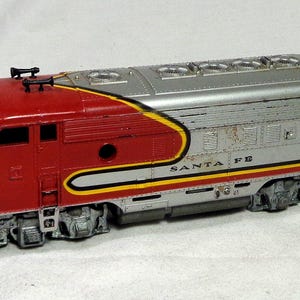 May include: A model train locomotive featuring a red and silver body with yellow accents. The front displays a yellow nose and the text SANTA FE is printed in black along the side. The engine includes silver wheels and detailed roof vents.