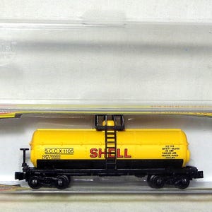 May include: A yellow and black Shell tanker train car model in a clear plastic case. The model has the Shell logo in red and black wheels. The case is open, showing the model inside.