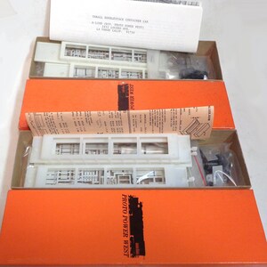May include: A model train kit in its original packaging. The kit is for a Thrall Doublestack Container Car, made by Proto Power West. The box is orange with white lettering and a picture of a train on it. The kit includes white plastic parts and instructions.