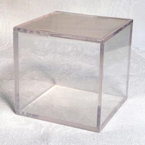 May include: A clear, transparent acrylic cube with a hinged top. The cube is empty and has a simple, minimalist design. The edges are neatly joined, creating a clean, modern aesthetic. Suitable for display or storage.