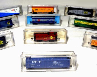 Puede incluir: Una colección de vagones de tren modelo en cajas de plástico transparente. Los vagones son de varios colores, incluyendo azul, naranja, rojo y verde, con marcas y logotipos detallados. Las cajas están dispuestas sobre una superficie blanca.