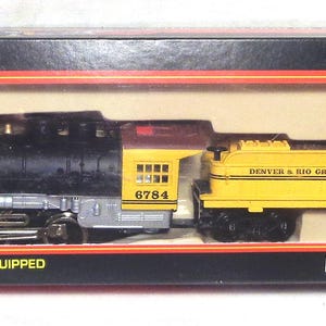 May include: Model Power HO scale locomotive in a box. The locomotive is black, silver, and yellow, with the text "DENVER & RIO GRANDE WESTERN" on the side. The box is black and red, with the text "HO SCALE" and "LOCOMOTIVE".