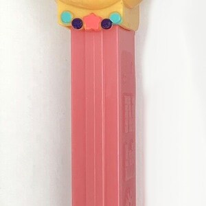 May include: A pink and yellow Sesame Street character with a pink hat and a big smile. The character is a toy dispenser with a pink plastic body.