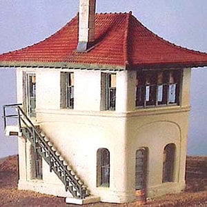 May include: A model railway signal box with a red tiled roof and a chimney. The building has white walls, arched windows, and a staircase. The model is set on a brown surface, typical of a railway scene.