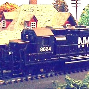 May include: A black model train engine with the letters "NW" on the side and the number "8824" printed on the front. The train is on a track in front of a model train station.