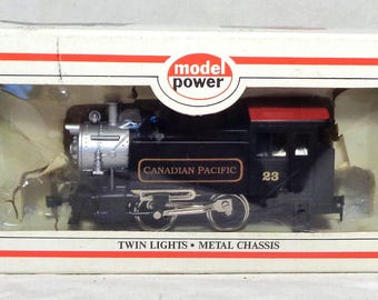 Op de afbeelding: Een zwarte en zilveren modeltreinlocomotief in een doos. De locomotief heeft de woorden "CANADIAN PACIFIC" en het nummer "23" op de zijkant. De doos leest "model power" en "TWIN LIGHTS • METAL CHASSIS".