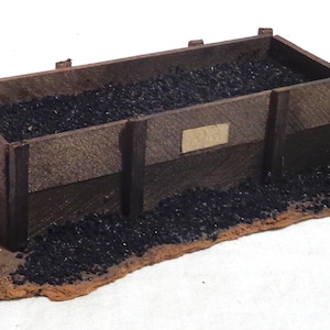Puede incluir: Un modelo en miniatura de un vagón de carbón de madera, lleno de material negro, descansa sobre una base marrón. El vagón tiene forma rectangular con soportes verticales y una pequeña etiqueta rectangular en el lateral. El modelo está diseñado para parecerse a un vagón de carbón.