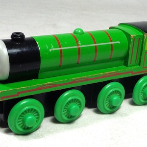 May include: A green wooden toy train engine featuring a black front, a grey face with a neutral expression, and four green wheels. The body is decorated with thin red horizontal lines and a black roof section on the rear cabin.
