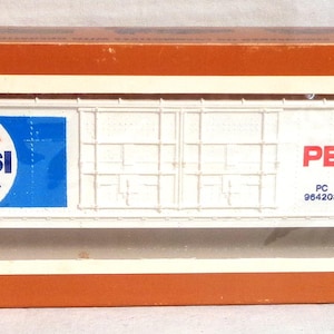 May include: A model train car in a box. The car is white with the Pepsi logo in red, white, and blue. The words "PEPSI-COLA" are printed in red. The box is orange with the "TYCO" logo.