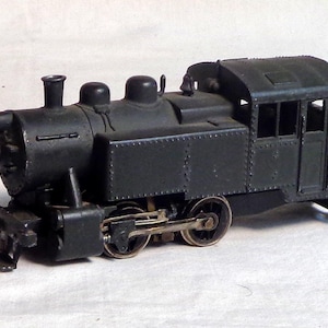 May include: A dark green toy train engine with a black cab and a single window. The engine has four wheels and a small, black smokestack.
