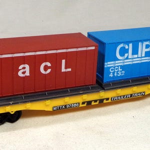 May include: A model flatcar train carriage in yellow with black wheels, carrying two shipping containers. One container is red with the white text 'a c L' and the other is blue with the white text 'CLIPPER' and 'CCL 4432'. The side of the flatcar features 'TRAILER TRAIN' and 'MTTX 97566'.