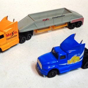 May include: Two toy semi-trucks, one orange and one blue. The orange truck is hauling a gray dump truck bed with the words "Maisto. Construction" printed on the side. The blue truck has a black trailer bed.