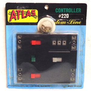 May include: A black Atlas Custom-Line #220 model train controller in a plastic package. The controller has two red and one green toggle switches, a black lever, and a gray slider. The package includes screws.