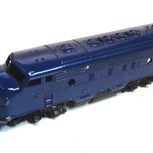 May include: A blue toy train engine with a detailed design. The engine has a rounded front and a long, rectangular body. The engine has several windows and a small door on the side.