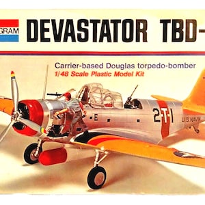 Puede incluir: Un kit de maqueta de plástico a escala 1/48 de un bombardero de torpedos Douglas TBD-1 Devastator. El modelo está pintado en un esquema de color plateado y amarillo con acentos rojos. El arte de la caja presenta una foto del modelo ensamblado con el texto "DEVASTATOR TBD-I" y "Torpedero Douglas basado en portaaviones" impreso en la caja.