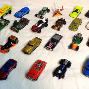 Könnte beinhalten: Eine Sammlung von 28 Spielzeugautos in verschiedenen Farben und Stilen, darunter ein rotes Rennauto, ein blauer LKW, ein grüner Monstertruck und ein gelber Sportwagen. Die Autos sind in Reihen auf einer weißen Oberfläche angeordnet.