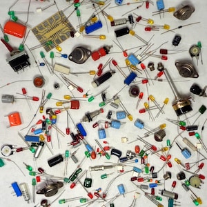 May include: A collection of assorted electronic components including various coloured LEDs, capacitors, resistors, transistors, a small printed circuit board, and a potentiometer with a metal shaft, all scattered across a plain white surface.