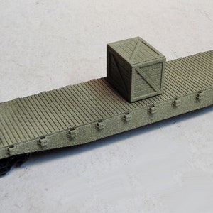 May include: A green model railroad flatcar with a wooden crate on top. The flatcar has metal rails and wheels. The crate is made of wood and has a wooden lid.