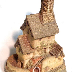 May include: A miniature ceramic model of a house with a tall chimney, a brown roof, and a winding path leading to the front door. The house is decorated with green ivy and red brick details.