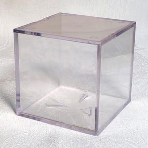 May include: A clear, square acrylic display case with a removable lid. The transparent box allows full visibility of the contents. The edges are slightly rounded. The box is on a white surface.