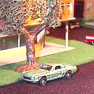 Może przedstawiać: Miniaturowa diorama przedstawiająca scenę miejską z zabawkami samochodami, budynkami i drzewem. Zielony klasyczny samochód zaparkowany jest na trawniku. Widoczny jest również czerwony samochód i żółty autobus szkolny.