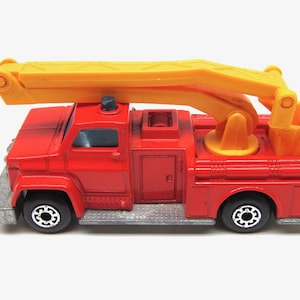 Puede incluir: Un camión de bomberos rojo de juguete con una escalera amarilla y una plataforma amarilla con un bombero de juguete en la parte superior. El camión tiene ruedas negras y un parachoques plateado.