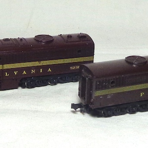 May include: Two maroon model train carriages with gold lettering reading "PENNSYLVANIA". The carriages are on a white surface. The train carriages have black wheels and are a vintage style.