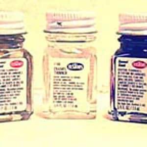 May include: Five small glass paint bottles with white lids are displayed. The bottles contain yellow, brown, clear, dark blue, and light blue paint. Each bottle has a label with text. The paint bottles are arranged in a row.