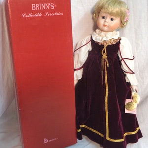 May include: A porcelain doll with blonde hair and blue eyes, wearing a burgundy velvet dress with gold trim and a white lace collar. The doll is holding a small tag that says "BRINN'S COLLECTIBLE EDITION". The doll is standing on a white surface and is next to a red box with the words "BRINN'S COLLECTIBLE PORCELAINS" printed on it.