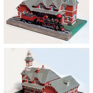 May include: A miniature red brick train station with a clock tower and a model train. The station has a gray roof and white trim. The train is black and red, with a coal car. The base is green and brown.