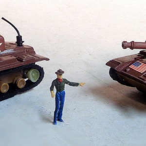 Puede incluir: Dos tanques de juguete marrones con el número 8127 en la torreta y una estrella blanca. Cada tanque tiene una pequeña calcomanía de la bandera estadounidense. Una figura en miniatura de una persona está de pie entre los tanques.