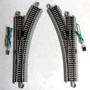 May include: Two grey plastic model railway track pieces with black rails. The pieces are curved and connect to form a points track. Each piece has a green wire attached.