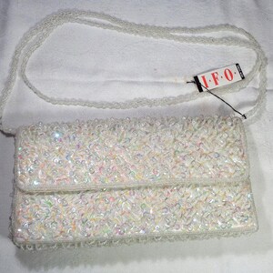 Puede incluir: Un bolso de mano blanco con cuentas y una correa de hombro a juego con cuentas. El bolso rectangular tiene un cierre de solapa y está cubierto de cuentas iridiscentes. Se adjunta una etiqueta con las letras "I.F.O.".