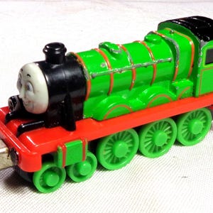 May include: A green toy steam locomotive with a black smokebox, a smiling face on the front, and a red chassis. The train features bright green wheels, a black roof, and silver-colored detailing along the boiler and cab sides.
