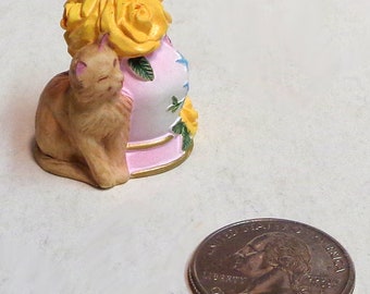 Puede incluir: Una figurita de cerámica de un gato sentado junto a un jarrón rosa y blanco con rosas amarillas. El jarrón tiene un borde dorado y un diseño floral.