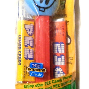 May include: Sealed PEZ candy dispenser package. The package is green with the PEZ logo and text. The dispenser is blue with a smiling face. Includes lemon candy and orange PEZ candy refills. The net weight is 1 oz.