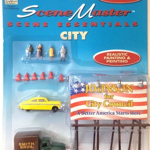 Op de afbeelding: SceneMaster City HO-schaalmodelset met miniatuurfiguren, verkeerskegels, een gele taxi, een groene vrachtwagen en een reclamebord met de tekst "JOHNSON for City Council". De verpakking is blauw met rode accenten.