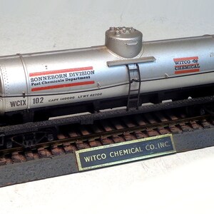 Pode incluir: Um vagão-tanque modelo de prata com rodas pretas e um chassi preto. O vagão-tanque tem letras vermelhas e brancas que dizem "SONNEBORN DIVISION Fuel Chemicals Department" e "WITCO CHEMICAL". O vagão-tanque está sentado sobre uma base marrom e cinza com uma placa dourada que diz "WITCO CHEMICAL CO., INC."